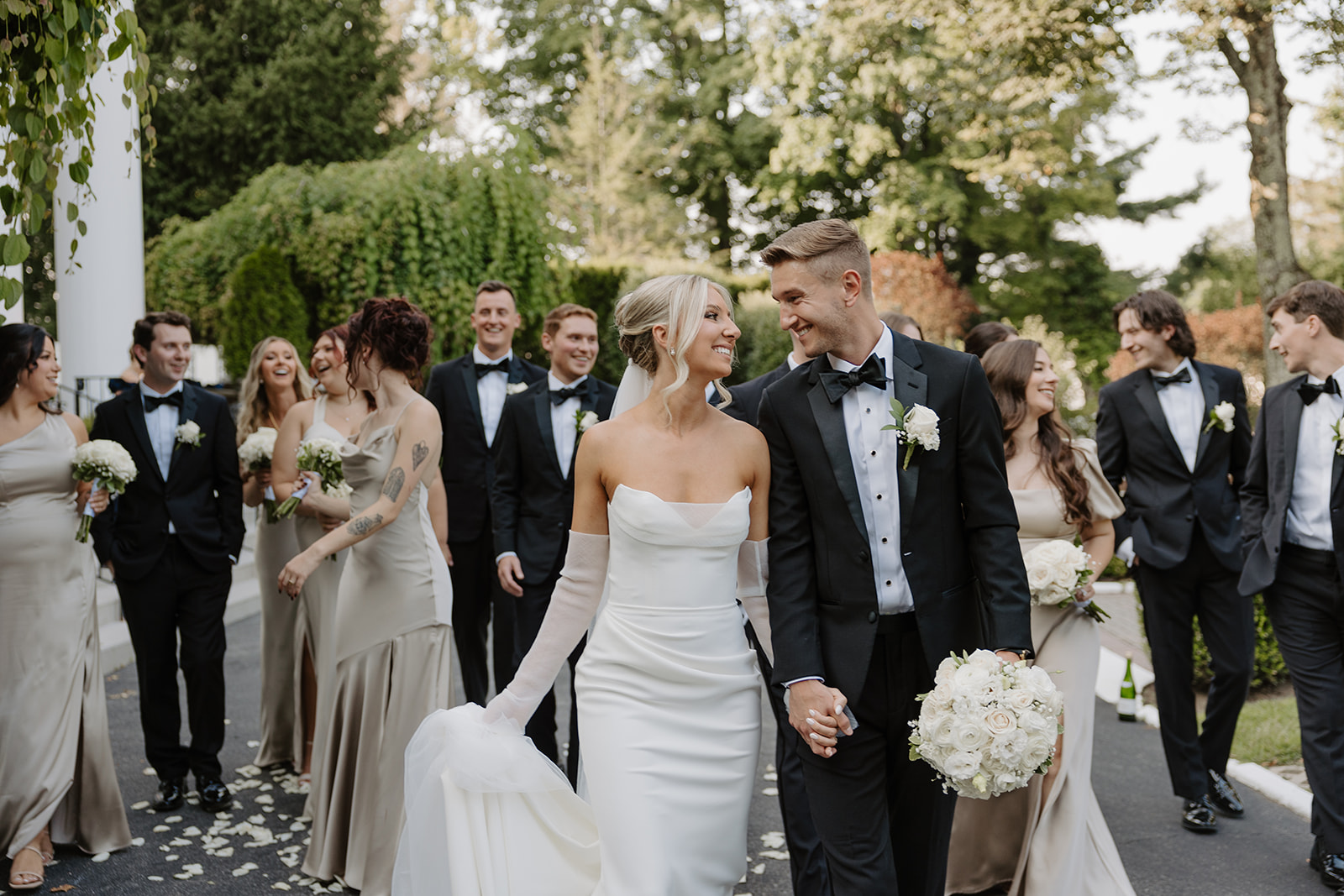Bride and groom lock eyes and smile while walking hand-in-hand, surrounded by their wedding party.